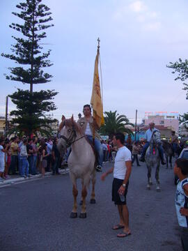 Φωτογραφία νεαρού καβαλάρη που προηγείται των υπολοίπων κουβαλώντας το αυθεντικό λάβαρο του Αγίου Γεωργίου. Μετά την εκκλησία του Αγίου Σπύριδων, οι αρματωμένοι και καβαλάρηδες ξεκινούν για το μοναστήρι, μεταφέροντας το λάβαρο ως φόρο τιμής.